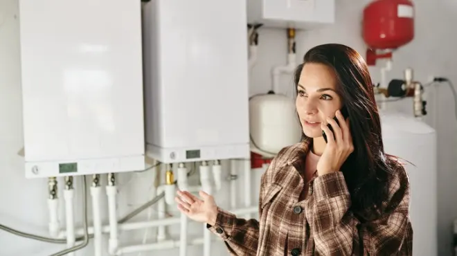 Kahverengi ekoseli ceketli kadın, beyaz kombilerin önünde telefonda konuşuyor. Arka planda beyaz bir su tankı ve kırmızı bir kazan var.