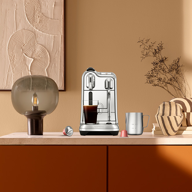 Kahverengi dolabın üstünde kahve makinesi, lamba, vazo ve kahve fincanı duruyor. Duvar bej renkte.