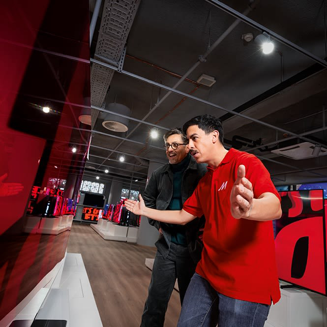 İki adam, kırmızı bir ekrana bakarken elektronik mağazasında duruyor. Öndeki adam kırmızı bir polo giyiyor.