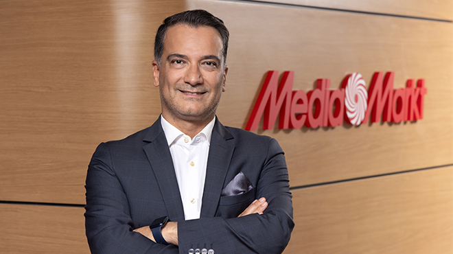 Takım elbiseli adam, MediaMarkt logosu önünde duruyor. Adamın kolunda saat var. Arka plan ahşap görünümlü.