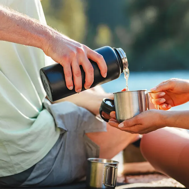 Açık havada, bir kişi termostan metal bir bardağa içecek dolduruyor. Bardak başka bir kişi tarafından tutuluyor.