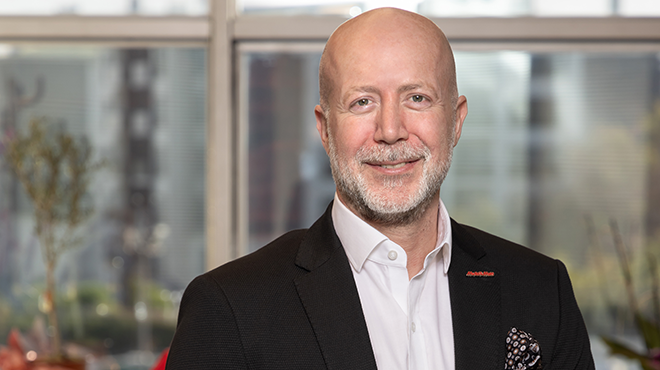 Kel sakallı, gülümseyen, takım elbiseli bir adam, pencere önünde duruyor.