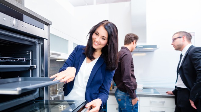 Mutfakta fırını inceleyen kadın, arka planda iki adam duruyor. Fırın metalik gri, mutfak beyaz renkte.