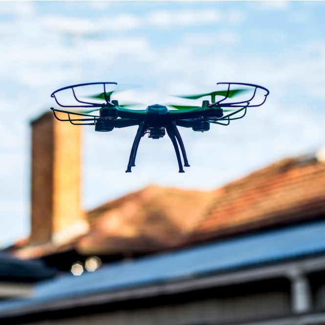 Mavi gökyüzünde, pervane korumalı, yeşil ve siyah drone uçuyor. Arka planda tuğla baca ve kiremitli çatı var.