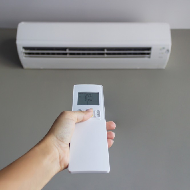 Duvara monte beyaz klima ve klimayı ayarlayan beyaz kumanda tutan bir el.