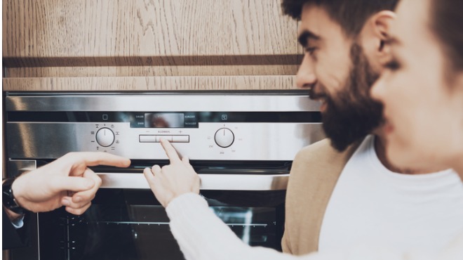 İki kişi fırını kullanıyor. Bir kişi düğmeye basıyor, diğeri izliyor. Arka planda ahşap dolap var.