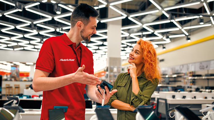 Bir elektronik mağazasında, kırmızı tişörtlü bir satıcı, turuncu saçlı bir müşteriye telefon gösteriyor.