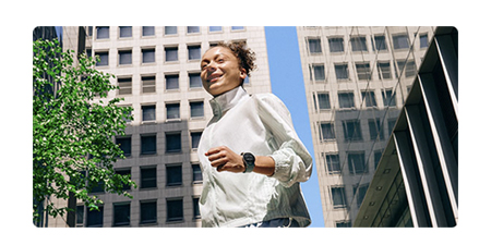 Şehirde koşan beyaz ceketli, akıllı saatli kadın. Arkada binalar ve ağaç var.