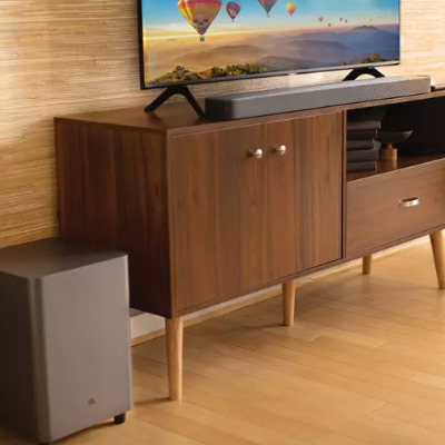 Kahverengi ahşap dolapta TV, ses çubuğu; yanda gri hoparlör. Duvar bej, zemin ahşap.