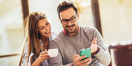 Gülümseyen bir kadın ve gözlüklü bir adam telefon ekranına bakıyor. Kadın elinde bir fincan tutuyor. Arka planda pencere var.