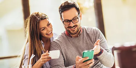 Gülümseyen bir kadın ve gözlüklü bir adam telefon ekranına bakıyor. Kadın elinde bir fincan tutuyor. Arka planda pencere var.