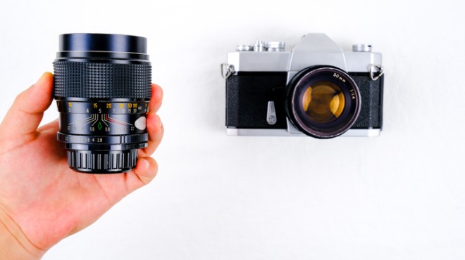 Beyaz fonda, bir elin tuttuğu siyah lens ve gümüş-siyah eski model bir fotoğraf makinesi görülüyor.