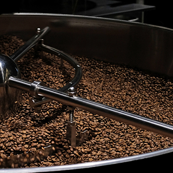 Kahve kavurma makinesinde kahverengi kahve çekirdekleri görülüyor. Metal karıştırma kolu ortada duruyor.