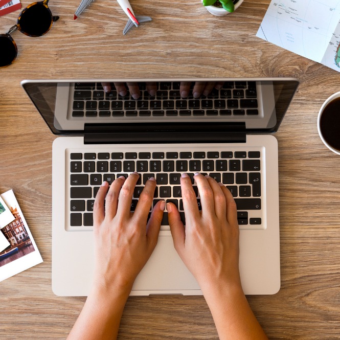 Tahta masada, elleriyle beyaz bir dizüstü bilgisayarda yazı yazan bir kişi görülüyor. Masada güneş gözlüğü, oyuncak uçaklar ve kahve var.